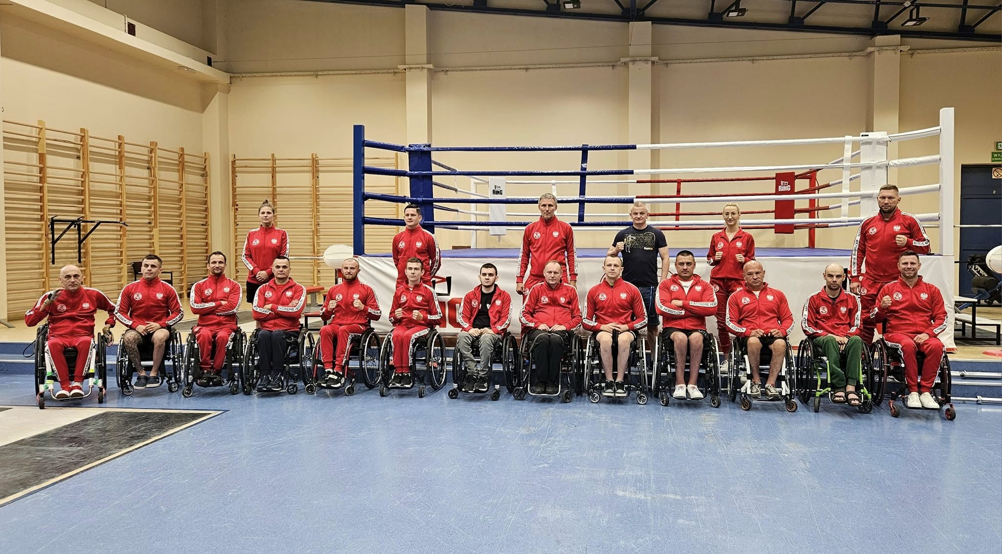 Grupowe zdjęcie reprezentacji Polski w paraboksie, składającej się ze sportowców na wózkach inwalidzkich i trenerów, stojących przed ringiem bokserskim w sali treningowej.