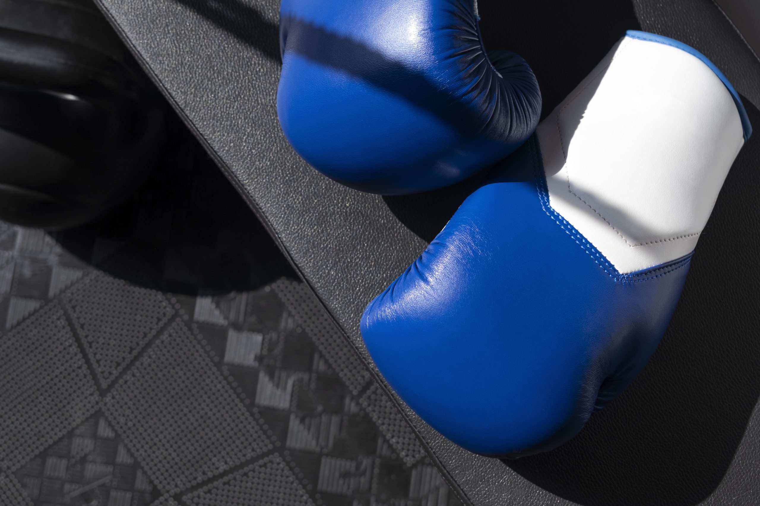 Zbliżenie na dwie niebiesko-białe rękawice bokserskie leżące na czarnej, gumowej macie treningowej. Obok leży duży, czarny odważnik kettlebell.